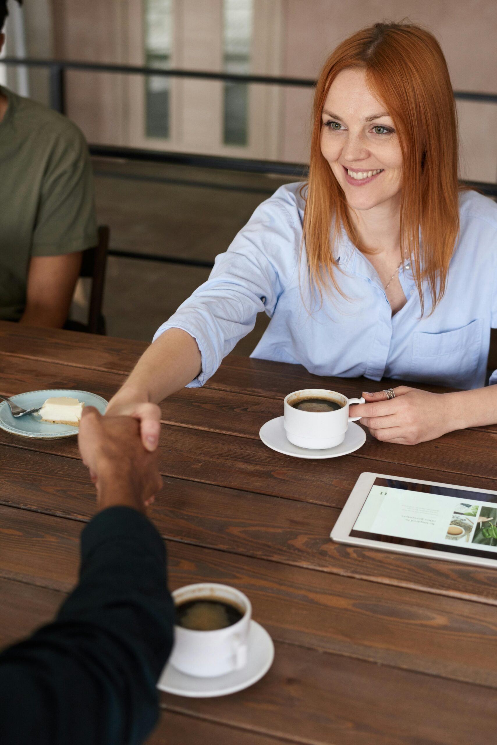 Smiling woman with red hair shakes hands across a table during a coffee meeting.
